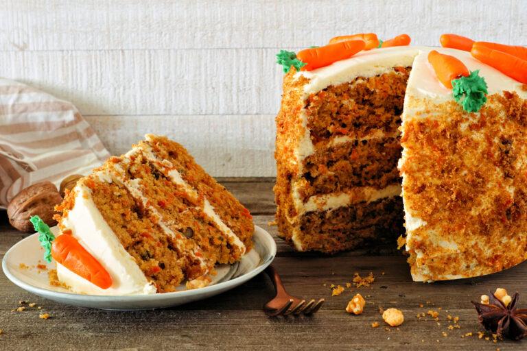 Slice of homemade carrot cake with cream cheese frosting and fondant carrots. Side view table scene with a white wood background.